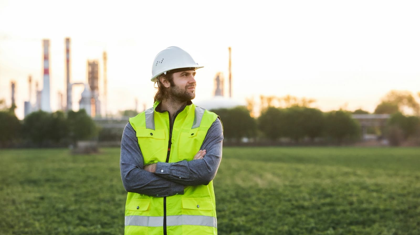 A worker in a hard hat and high-visibility vest standing in a field near an industrial plant